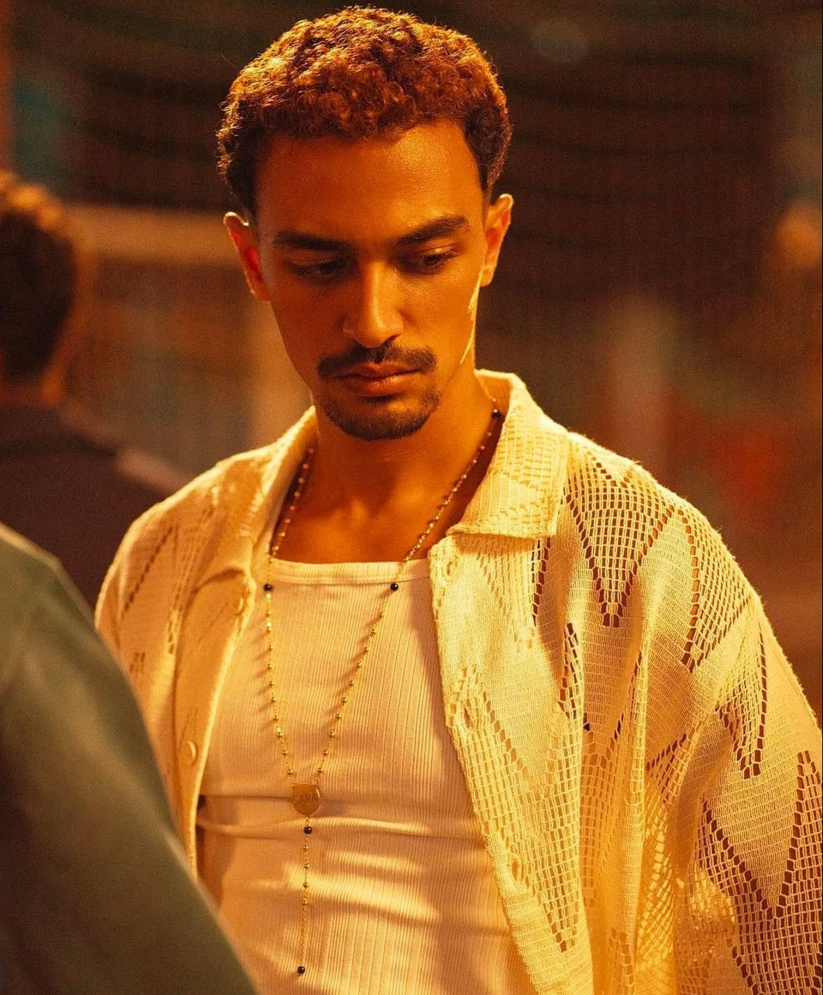 Man wearing a patterned beige shirt with a blurred background and silver necklace with beads and engraved name on it

